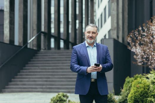 An older gray-haired man, a businessman in a suit, is traveling to another city, to another country. He is holding the phone in his hands, looking for directions on GPS. Walks near a modern building.