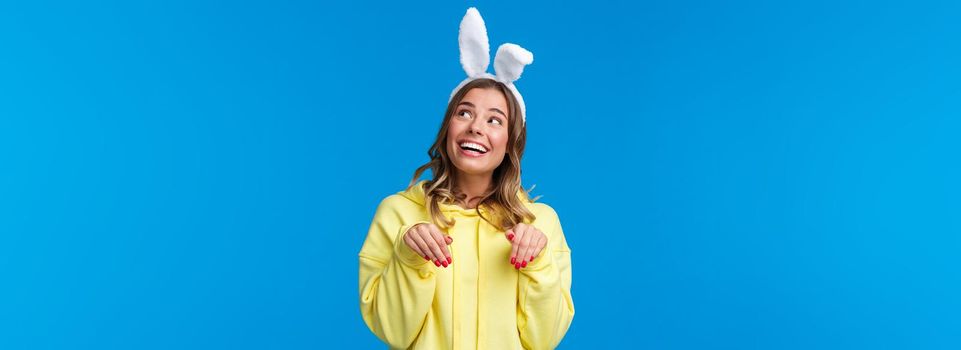 Holidays, traditions and celebration concept. Dreamy and cute young woman smiling lovely with rabbit ears, look upbeat upper right corner grinning make paws gesture, blue background.