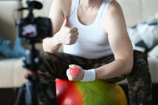 Athlete man holds thumbs up and raises dumbbells sitting on ball at home in front of camera. Recommendations for home fitness and sports training remotely