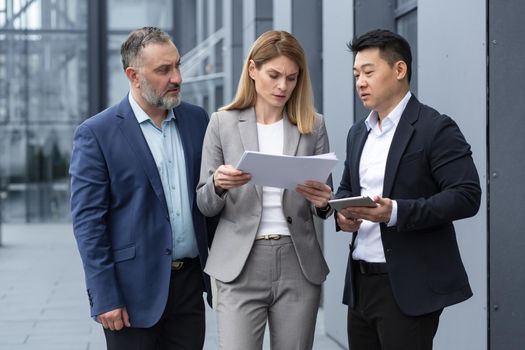 Diverse business team three colleagues employees discuss documents contract outside office building businessman and businesswoman holding bills and financial reports talking