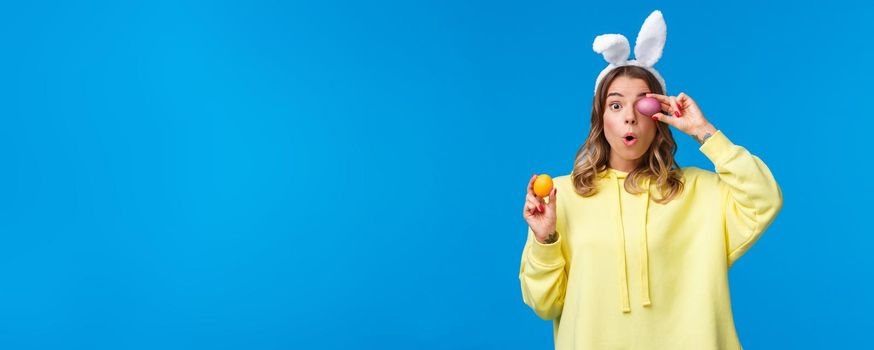 Holidays, traditions and celebration concept. Amused lovely european female blonde in rabbit ears, look excited and surprised as holding two painted Easter day eggs, blue background.
