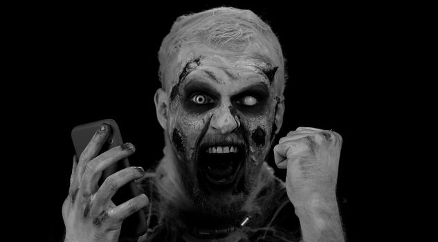 Happy excited sinister man in carnival costume of Halloween crazy zombie looking smartphone display sincerely rejoicing win, receiving message with good news, success luck. Cosplay wounded undead