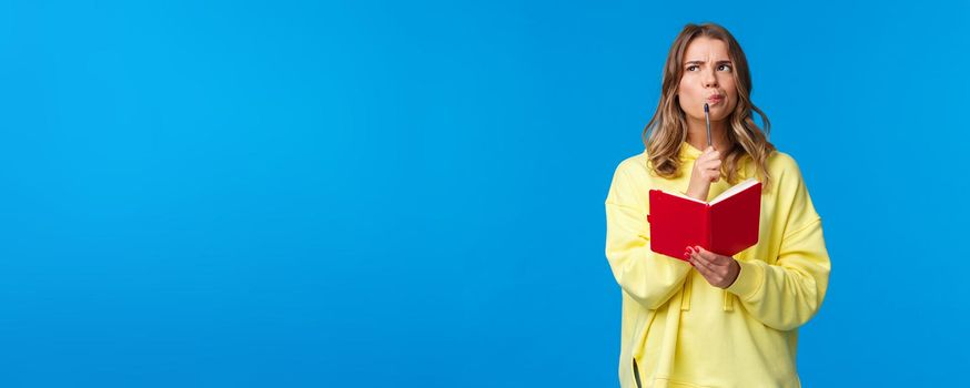 Thoughtful blond european female making plans, touching lip with pen and looking up pondering while thinking taking notes, holding red notebook, standing blue background. Copy space