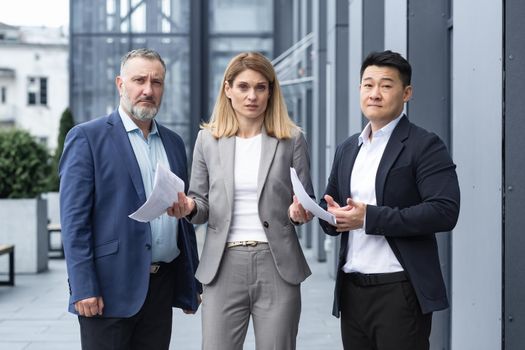 Frustrated female business boss with documents looking at camera, team of diverse colleagues outside office building upset with result of work report