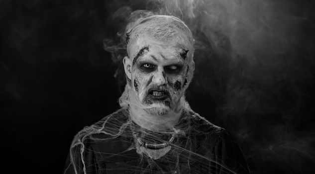 Sinister man with horrible scary Halloween zombie makeup in convulsions making faces, looking ominous, trying to scare. Dead guy with wounded bloody scars face isolated against black wall background