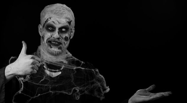 Creepy man with bloody scars face, Halloween stylish zombie make-up. Scary wounded undead guy showing thumbs up and pointing at left on blank space. Place for your advertisement logo. Sinister monster