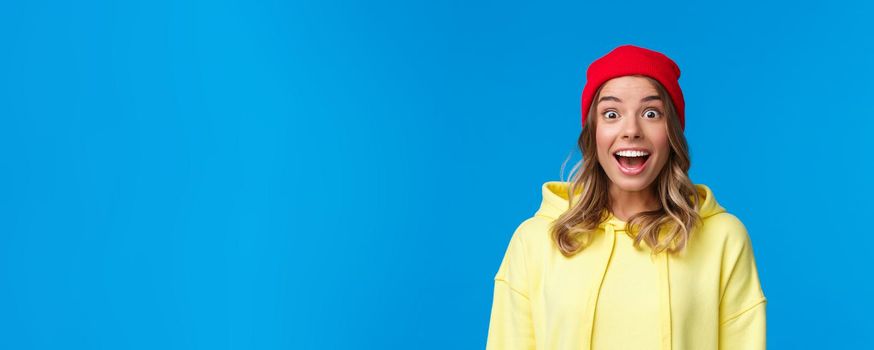 Close-up portrait of excited and intrigued young hipster girl in red beanie and yellow hoodie, gasping amazed look camera wondered and entertained, standing blue background.