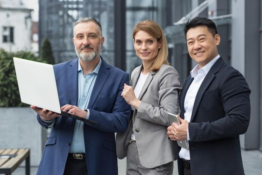 Portrait dream team IT specialists professionals team leaders outside office building looking at camera holding laptop discussing successful application project, specialists business suits smiling.