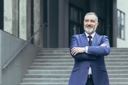 Portrait of senior handsome real estate agent man. He stands next to a business suit near a modern building, looks to the side, smiles