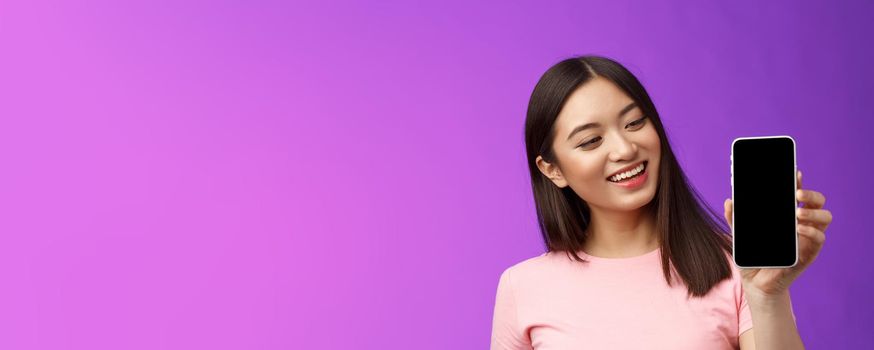 Glad cheerful good-looking young asian woman smiling satisfied hold smartphone, show phone screen look phone display pleased, introduce app, present application, purple background.