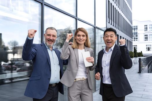 Successful diverse business team, three loving men and woman looking at camera and happy celebrating victory, team dreaming outside office building, business group in business suits