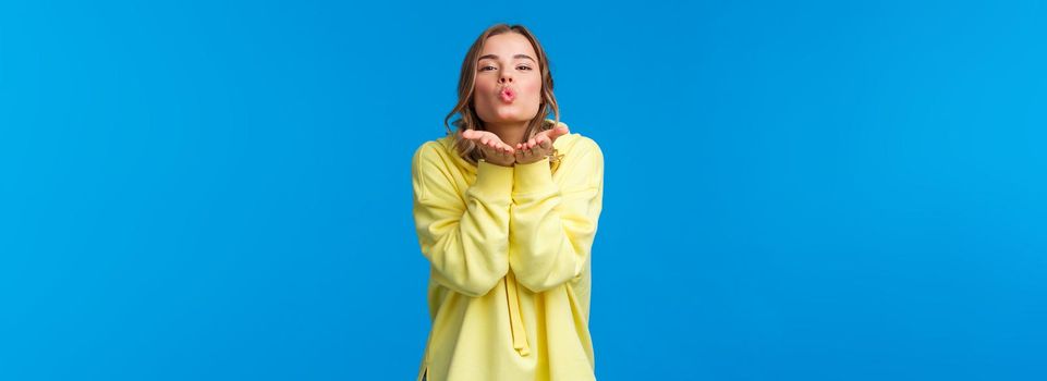 Send you passionate kisses through camera. Tender and silly girlfriend with blond short hair holding palms near folded lips and make mwah gesture, standing blue background.