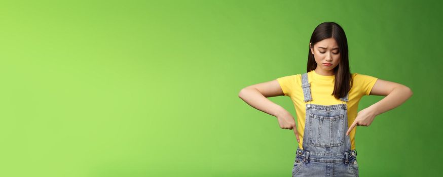 Gloomy disappointed cute innocent asian teenage girl look down upset, frowning look pointing down sorrow, unhappy terrible situation happened, express sadness and distress, green background.