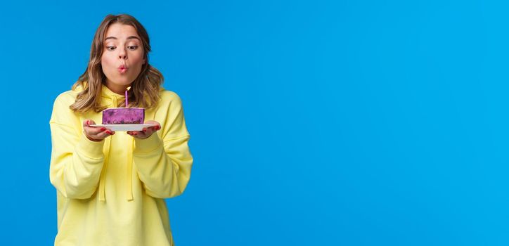 Celebration, party and lifestyle concept. Silly cute european blond woman in yellow hoodie, making wish holding fruit cake blowing out b-day candle, standing blue background at birthday party.