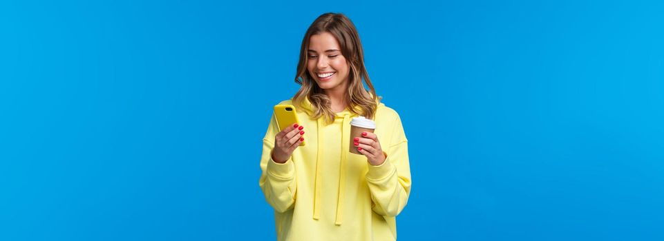 Carefree joyful european female in yellow hoodie, holding take-away coffee and browsing internet in mobile phone, texting friend laughing over funny video or meme, blue background.