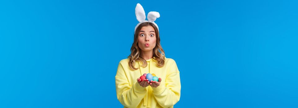 Holidays, traditions and celebration concept. Silly cute caucasian blond girl present you Easter eggs, painted it for holiday, show mwah kiss expression and wear rabbit ears, blue background.