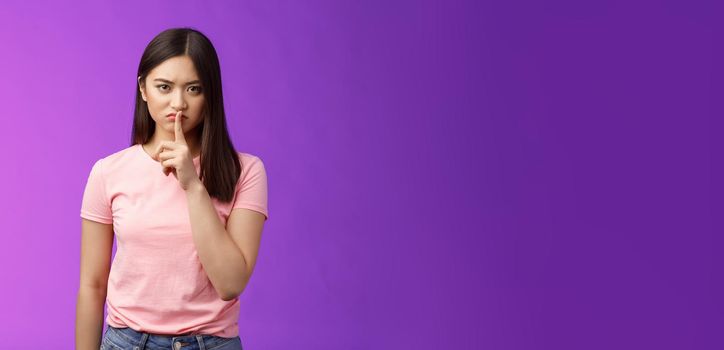 Shut up, seriously. Angry unsatisfied asian woman brunette frowning make hush sign, hold index finger on lips grimacing annoyed, prohibit speak, shushing stay silent, stand purple background.