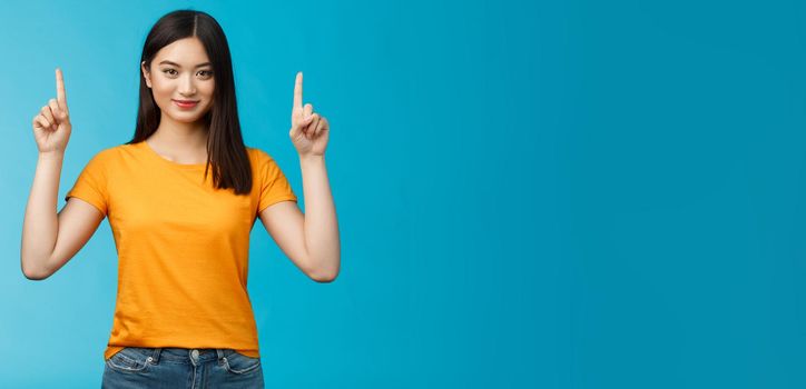 Heads up see awesome promo. Assertive good-looking confident asian woman showing friend advertisement, raise index fingers pointing top, smiling camera self-assured, stand blue background determined.