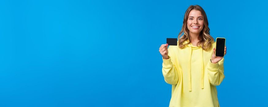 Banking and finance concept. Portrait of happy smiling blond young girl using credit card, showing it and smartphone display, satisfied with new application helps shopping online, blue background.