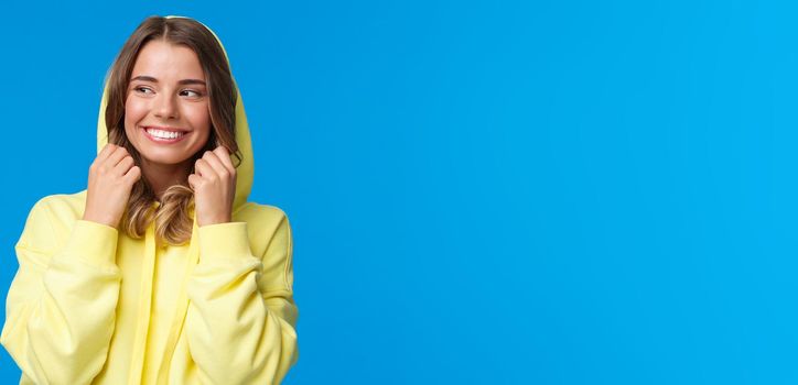 Close-up portrait beautiful charming blond girl put on hood of sweatshirt and smiling, look away, feel chilly as walking on spring day, standing carefree over blue background. Copy space