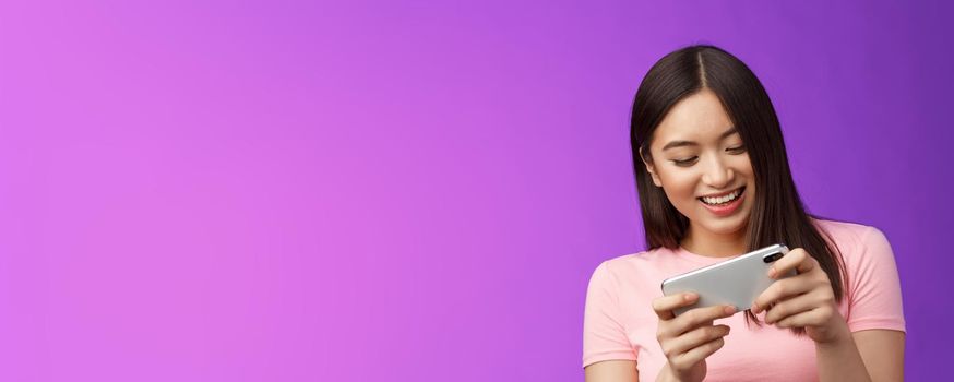 Close-up joyful attractive asian woman brunette having fun spend time playing smartphone game, laughing smiling eager win race, hold phone horizontal beating score, purple background.