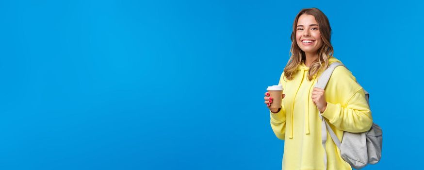 Joyful young blond female student with backpack smiling camera satisfied as holding cup of take-away coffee from favorite cafe after classes, drinking beverage, stand blue background.