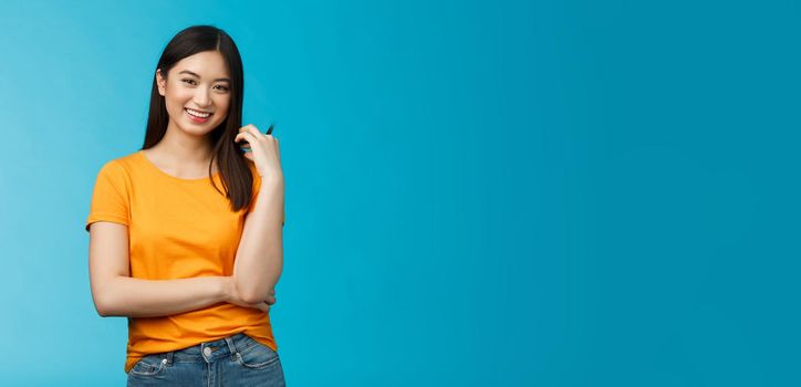 Cheerful asian girl with dark short haircut touching hair strand smiling lively, cross one arm chest, laughing, carefree talking casually friends, discuss university lifestyle, blue background.