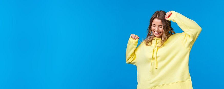 Happy and relaxed, carefree caucasian blond girl having fun at party, listening music vibing, raise hands up and dancing joyful, smiling with closed eyes, standing blue background.