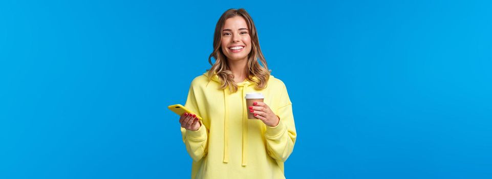 Cheerful young female student standing near cafe ordered take-away coffee, drinking from paper cup and holding mobile phone, look at camera with beaming white smile, satisfied.