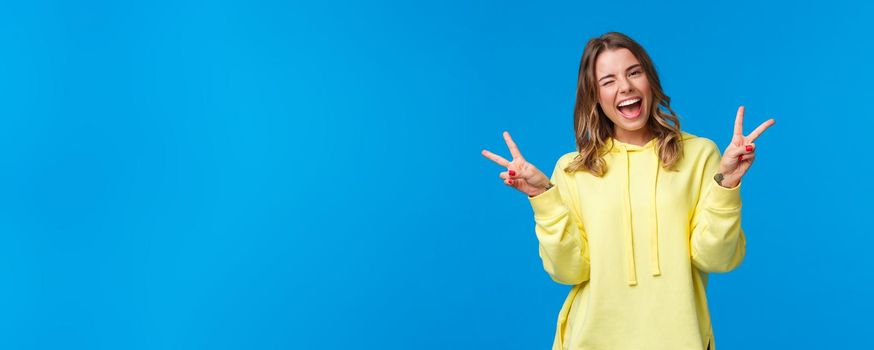 Lets have some fun. Carefree upbeat friendly-looking kawaii blond girl in yellow hoodie, wink and smiling white teeth as showing cute peace gestures, standing blue background.