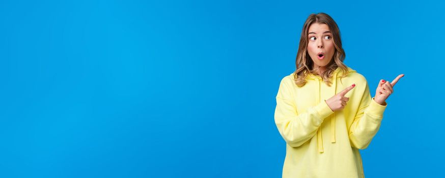 Wow something interesting. Intrigued and surprised alluring blond woman in yellow hoodie talking about last rumors, pointing and looking right with impressed expression, blue background.