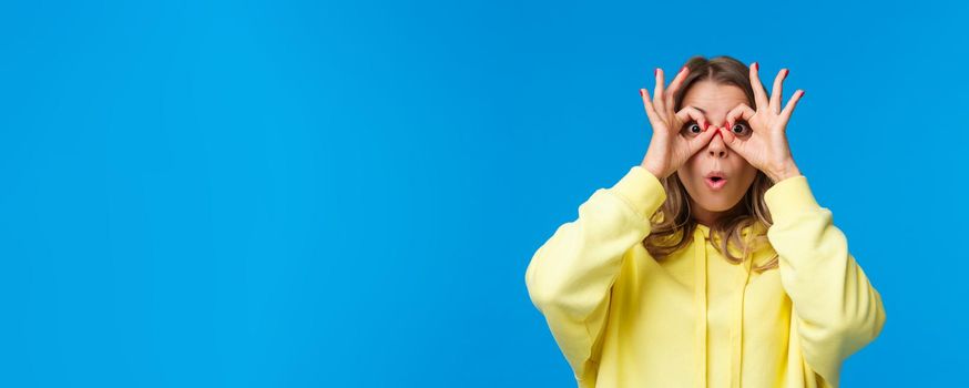 Impressed and intrigued, curious young caucasian girl in yellow hoodie looking at something awesome, found great product special discount, make glasses with fingers on eyes, blue background.