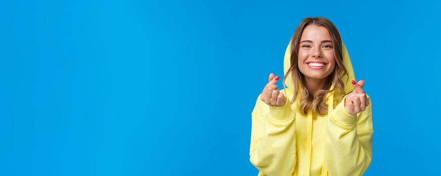 Close-up portrait of cute and silly blond caucasian girl in yellow hoodie, showing korean heart gesture and smiling, vibing, enjoying party, having fun over blue background. Copy space