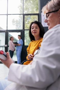 Senior doctor measuring hypertension and blood pressure to do heart exam with tonometer in hospital waiting area. Medic consulting asian patient with cardiology instrument, medicine support