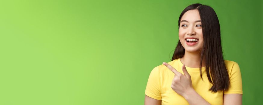 Close-up cheerful friendly asian outgoing girl having fun, pointing turn left amused, lively talking, discuss friends interesting event, stand green background chatting carefree entertained.