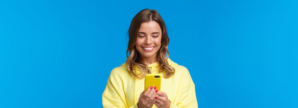 Close-up portrait silly beautiful blond european woman in yellow hoodie, smiling at mobile phone display as watching lovely video, messaging with boyfriend, standing blue background.