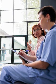 Woman nurse consulting asian patient in hospital lobby, filling medical report papers discussing treatment. Assistant doing checkup examination with senior woman in waiting area.