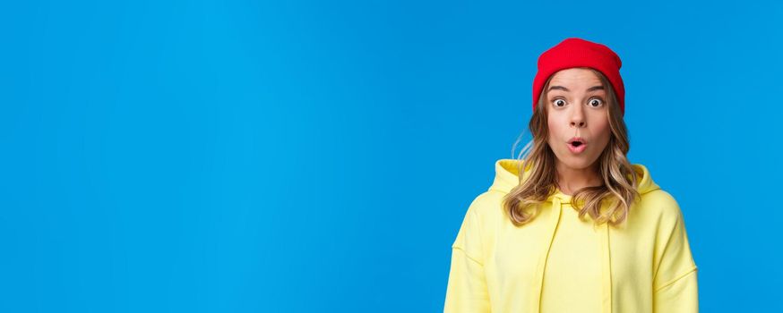 Wow tell me more. Intrigued and curious blond european girl staring astonished, gasping impressed, watching something exciting, hear interesting news, standing in red beanie and yellow hoodie.