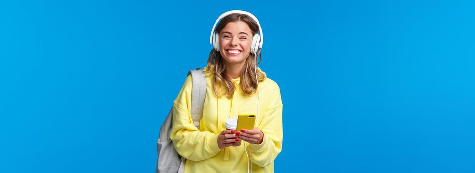 Cheerful pretty young female student with backpack, listening favorite playlist in headphones, laughing and smiling at camera carefree, hold mobile phone, stand blue background.