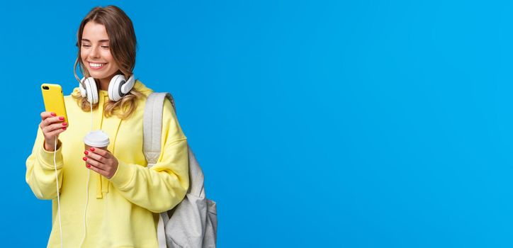 Girl using mobile phone to contact friend after college, texting as walking along street with backpack, take-away coffee cup and headphones, smiling joyfully smartphone display.