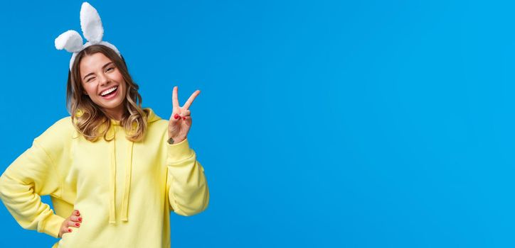 Holidays, traditions and celebration concept. Lovely and tender blond girl in yellow hoodie, tilt head kawaii wink and smiling as wearing rabbit ears, show peace gesture for goodwill, happy easter.