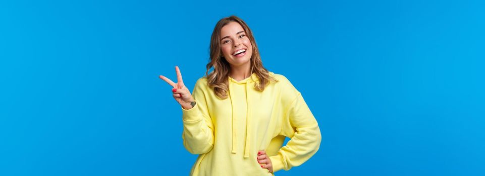 Staying bright sie. Carefree happy young silly blond girl in yellow hoodie sending positive vibes, show kawaii peace sign and smiling with white teeth, standing blue background.