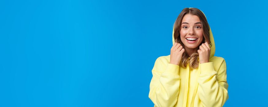 Lifestyle, people and youth concept. Close-up portrait of beautiful smiling blond female put on hoodie, looking camera with upbeat enthusiastic expression, standing blue background.