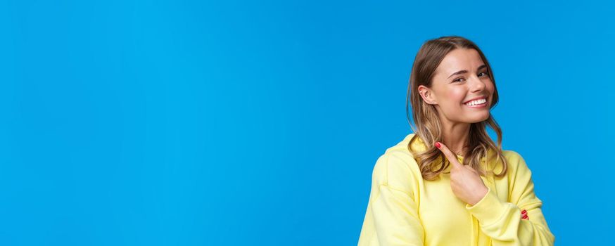 Close-up portrait of beautiful blond girl with short hair pointing at her piercing in ear, smiling proud and satisfied, change her look, standing blue background in yellow hoodie. Copy space