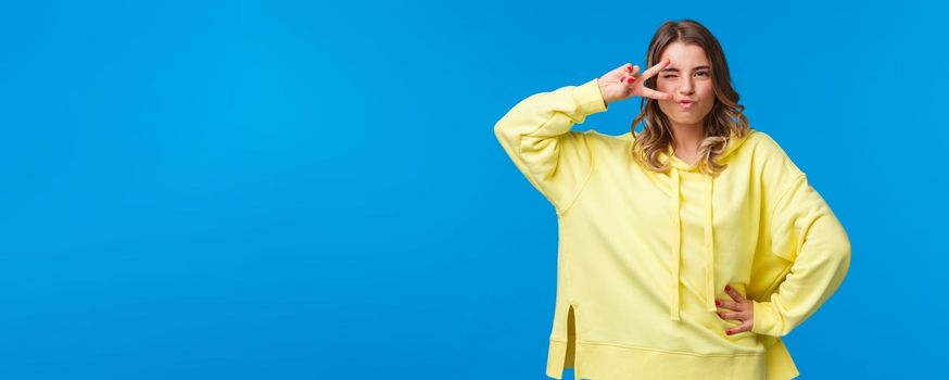 Portrait of sassy alluring blond caucasian woman dancing disco, show peace sign near eye and looking daring and sexy camera, standing in assured pose over blue background.