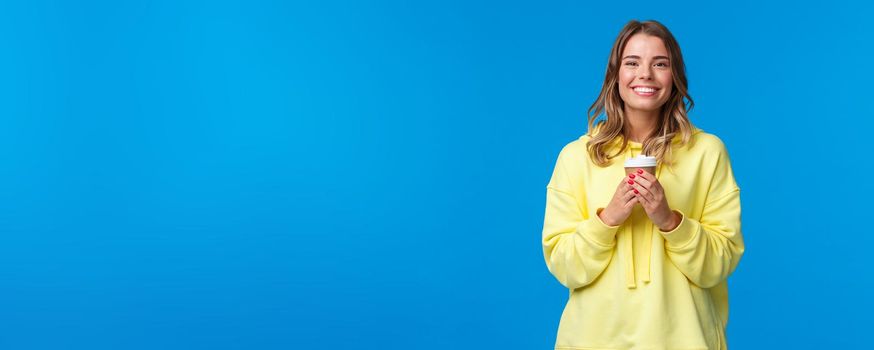 Girl enjoying tasty cup of morning coffee from favorite cafe. Cheerful lovely blond woman in yellow hoodie smiling white teeth and holding paper cup of take-away tea, stand blue background.