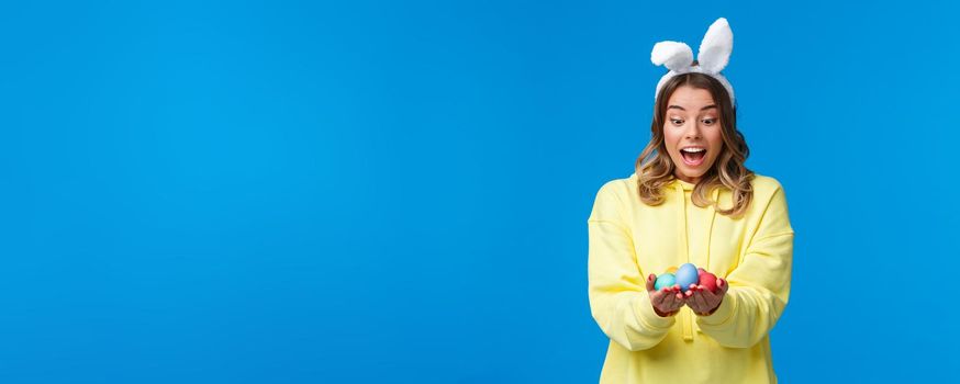 Surprised emotive cute blond caucasian girl playing holiday game, found Easter eggs look amused, wearing rabbit ears as celebrating religious holiday on sunday, blue background.