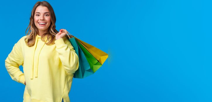 Shopping time. Girl relaxing after hard working days buying things in store on weekend, hold bags on back and smiling relieved, release stress through purchasing clothes, blue background.