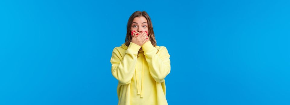 Shocked and speechless impressed blond woman close mouth with hands and stare astonished, hear shook rumour, gossiping make astounded face, standing blue background.