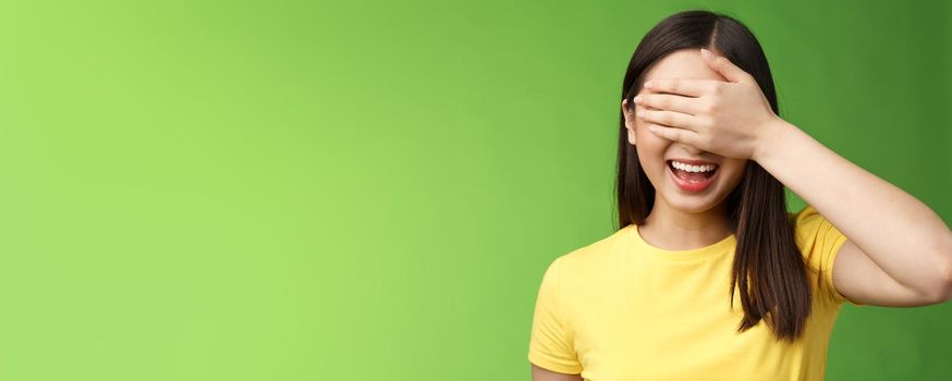 Lively joyful friendly-looking asian girl shut eyes palm, laughing happily, standing amused anticiapting b-day gift, counting ten, playing hide-n-seek, stand green background entertained.